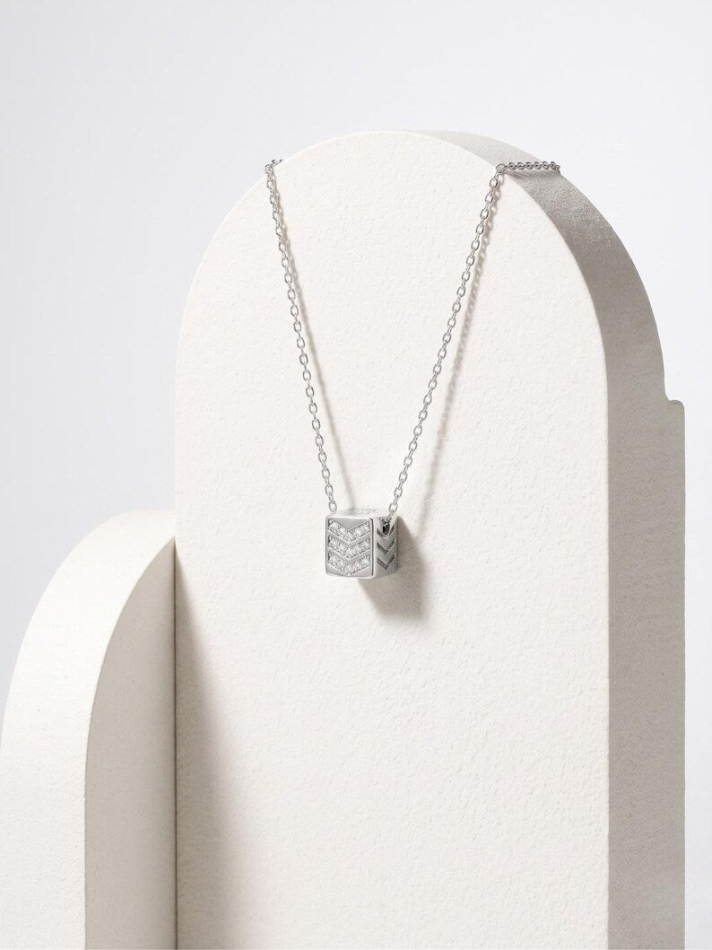 A silver pendant necklace with a textured, geometric cube charm displayed on a white arched jewelry stand against a light gray background.