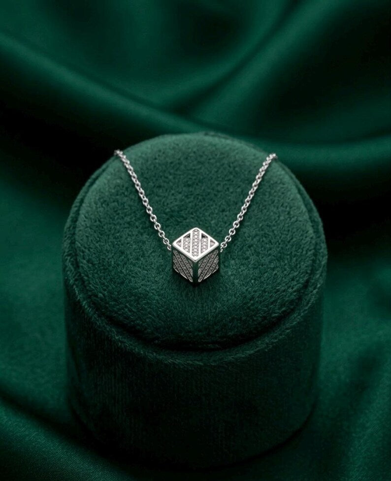 A silver geometric pendant necklace with a textured pavé-style design on a delicate chain, displayed on a dark green velvet jewelry stand against a matching green satin background.