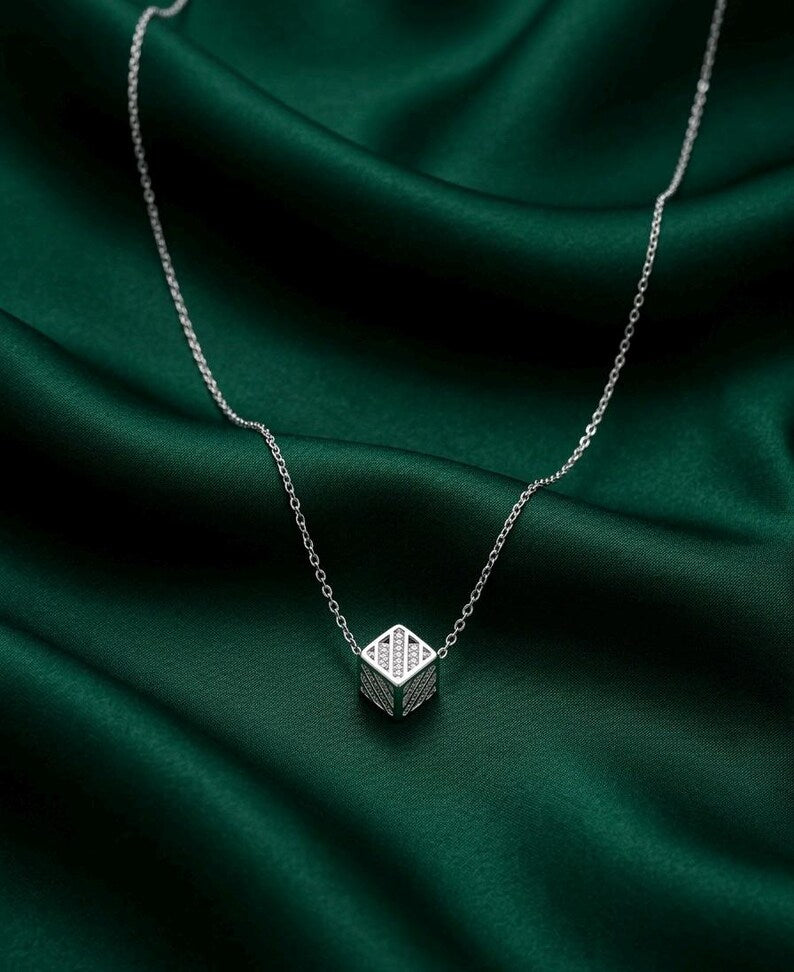 A silver chain necklace with a small geometric cube pendant featuring a textured grid pattern, displayed on draped dark green satin fabric.
