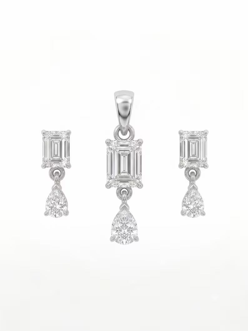A jewelry set featuring a central pendant and two matching drop earrings, each with an emerald-cut diamond and a pear-cut diamond, set in a silver-toned metal on a plain white background.