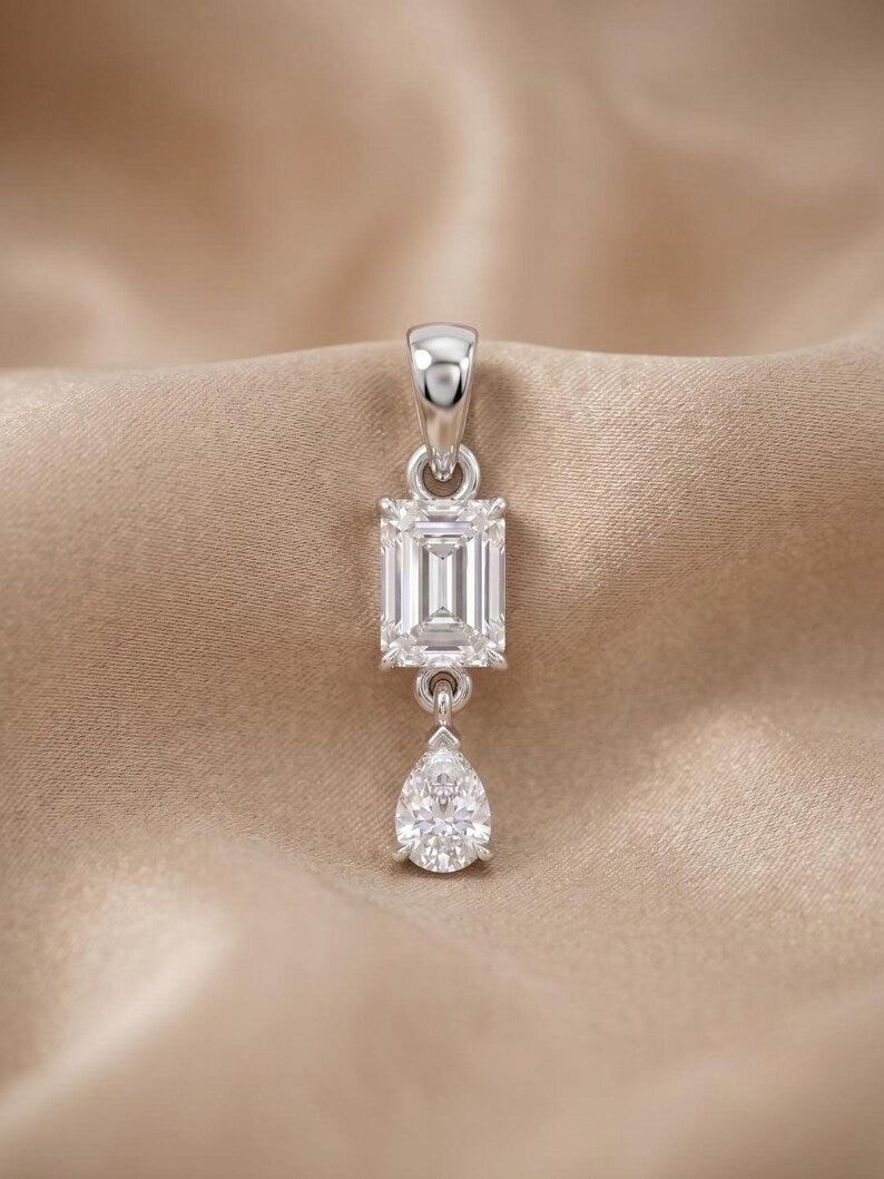 A silver pendant featuring an emerald-cut clear gemstone and a dangling pear-cut clear gemstone, displayed on a beige satin fabric background.