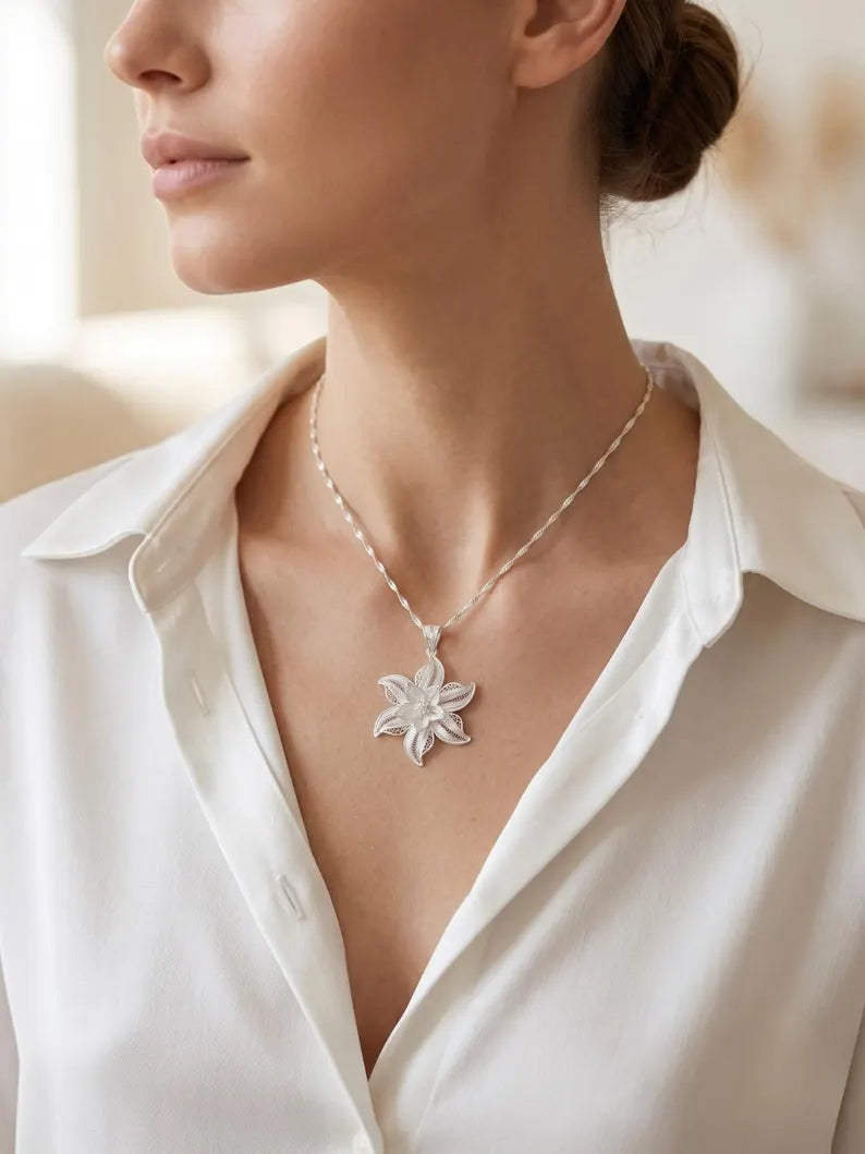 A close-up of a woman wearing a silver-toned flower-shaped pendant necklace with a delicate beaded chain, paired with a white collared blouse.