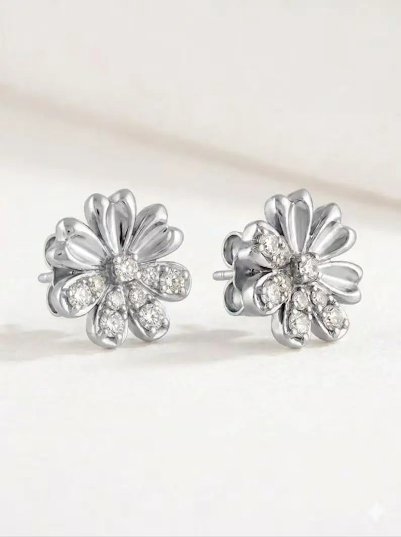 A pair of silver flower-shaped stud earrings with clear gemstones, displayed on a clean, light beige background.