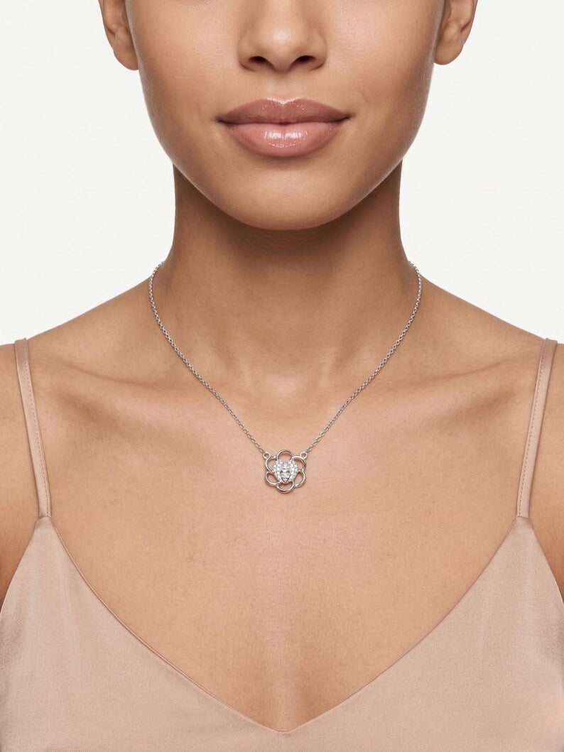 A close-up of a model wearing a delicate silver chain necklace with a flower-shaped pendant featuring a clear central gemstone and silver petals, paired with a beige camisole top.