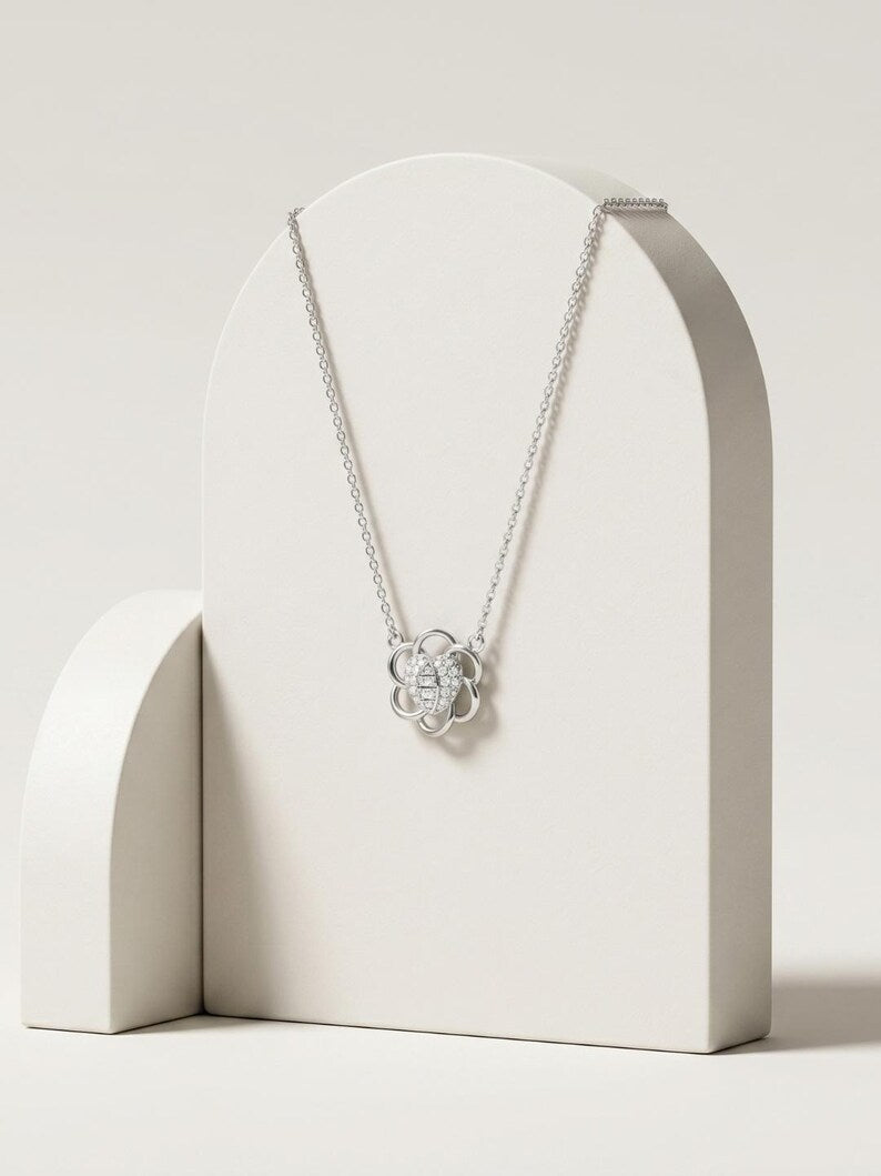 A silver pendant necklace with a flower-like design and central letter 'S', displayed on a white arched stand against a warm off-white background.