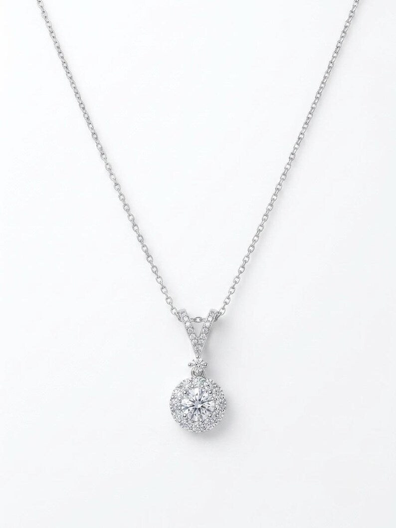 A silver-toned necklace featuring a round halo pendant set with clear gemstones, suspended from a delicate chain against a plain white background.