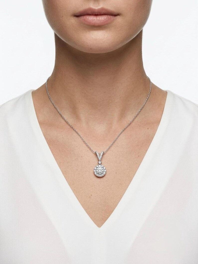 A close-up of a model wearing a delicate silver chain necklace with a round, pavé-set pendant on a white V-neck top against a clean white background.