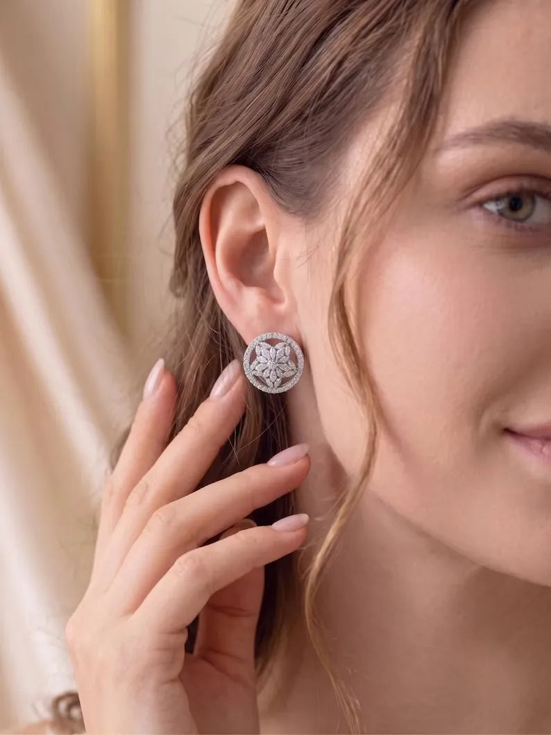A close-up lifestyle shot of a woman wearing a round silver-toned Marquise halo stud earrings presented with a detailed floral or starburst crystal design, with her hand gently touching her hair.