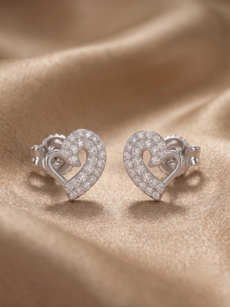 A pair of silver heart-shaped stud earrings paved with clear stones, displayed on a draped beige satin fabric background.