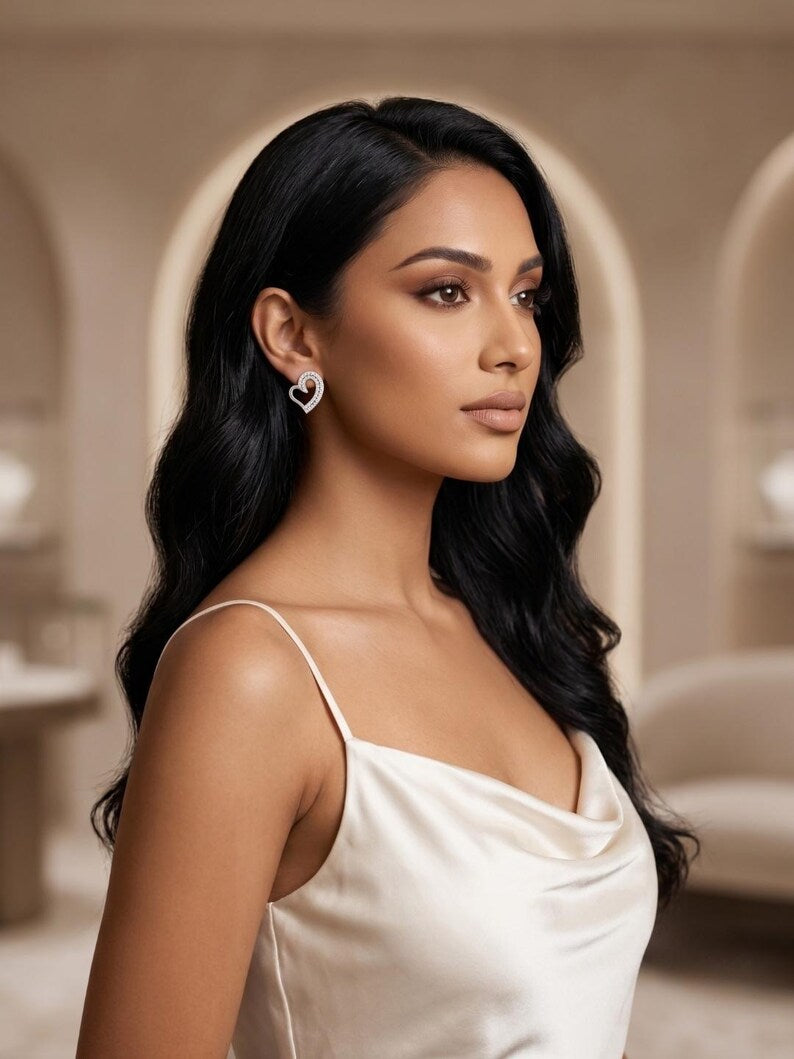 A woman with long, wavy black hair is shown in profile wearing a white satin camisole and a small silver heart-shaped stud earring in a softly lit indoor setting with beige arches.