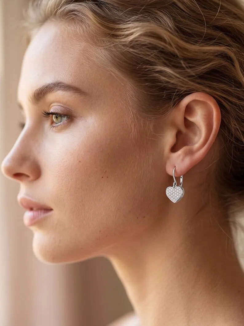 A close-up side profile of a woman wearing a small silver heart-shaped hoop earring with a textured pavé-style heart dangle.