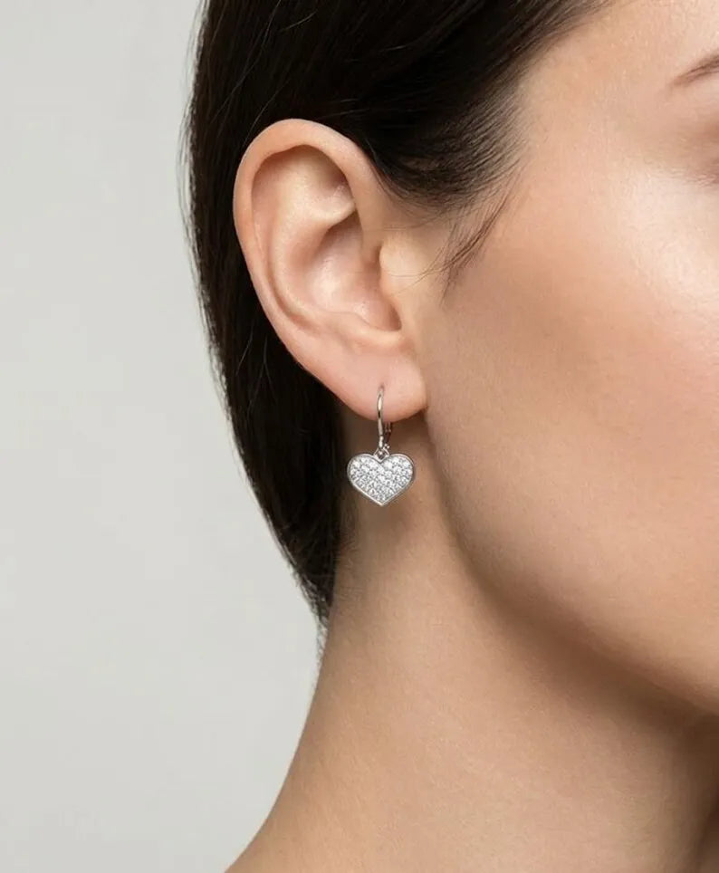 A close-up of a model's ear wearing a small silver heart-shaped dangle earring paved with clear stones, against a light gray background.