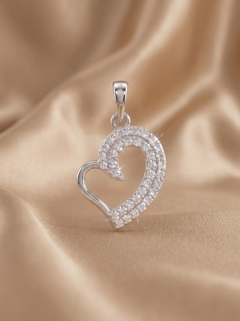 A silver heart-shaped pendant adorned with pavé-set clear stones, displayed on a draped beige satin fabric background.