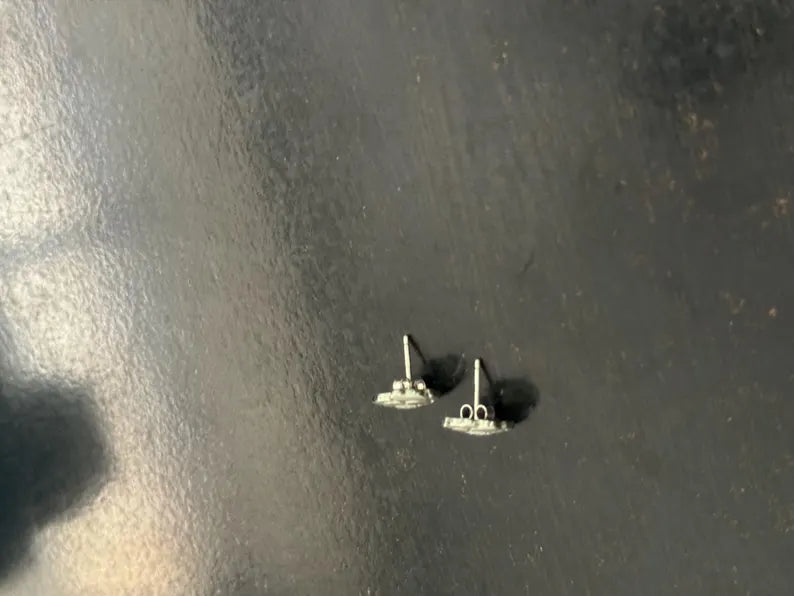A pair of small silver stud earrings with a circular disc design, shown on a dark, textured surface.