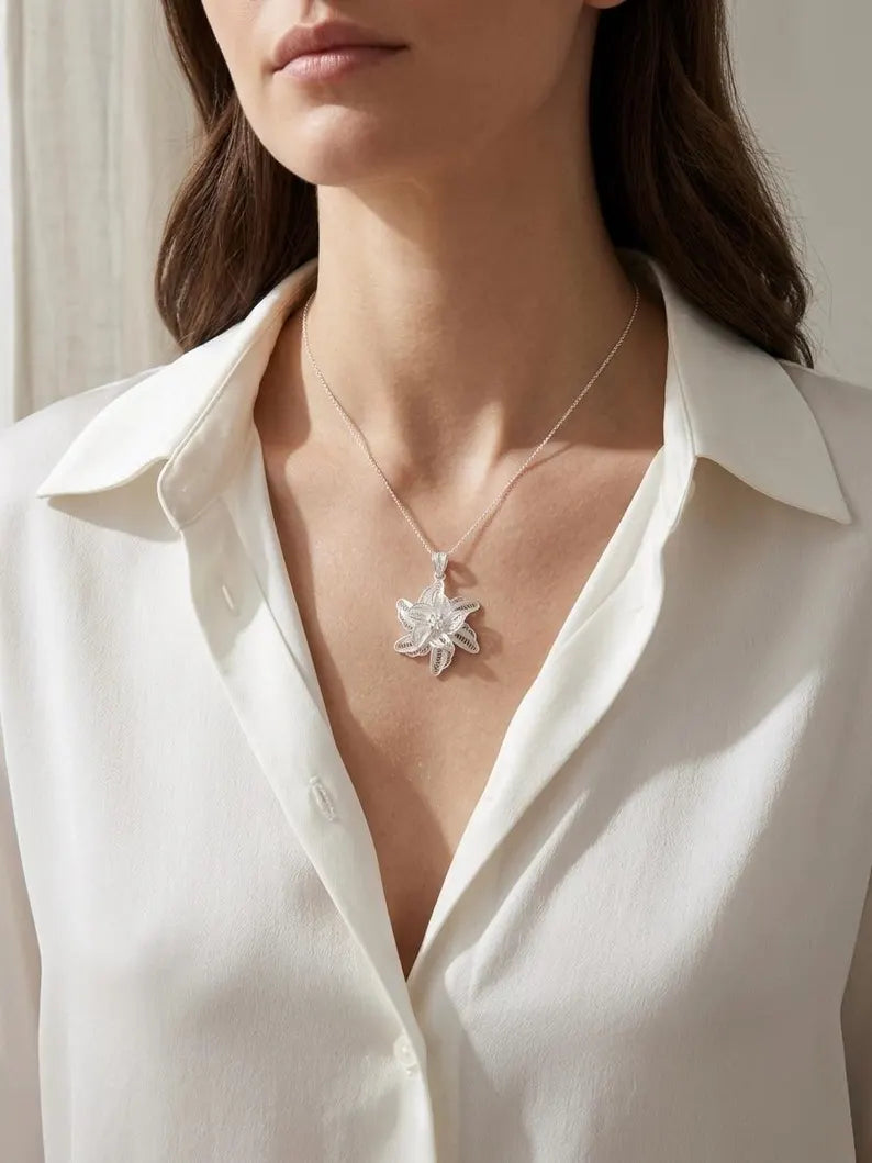 A close-up of a woman wearing a delicate silver pendant necklace with a starburst or snowflake-like design, styled with a white collared blouse.