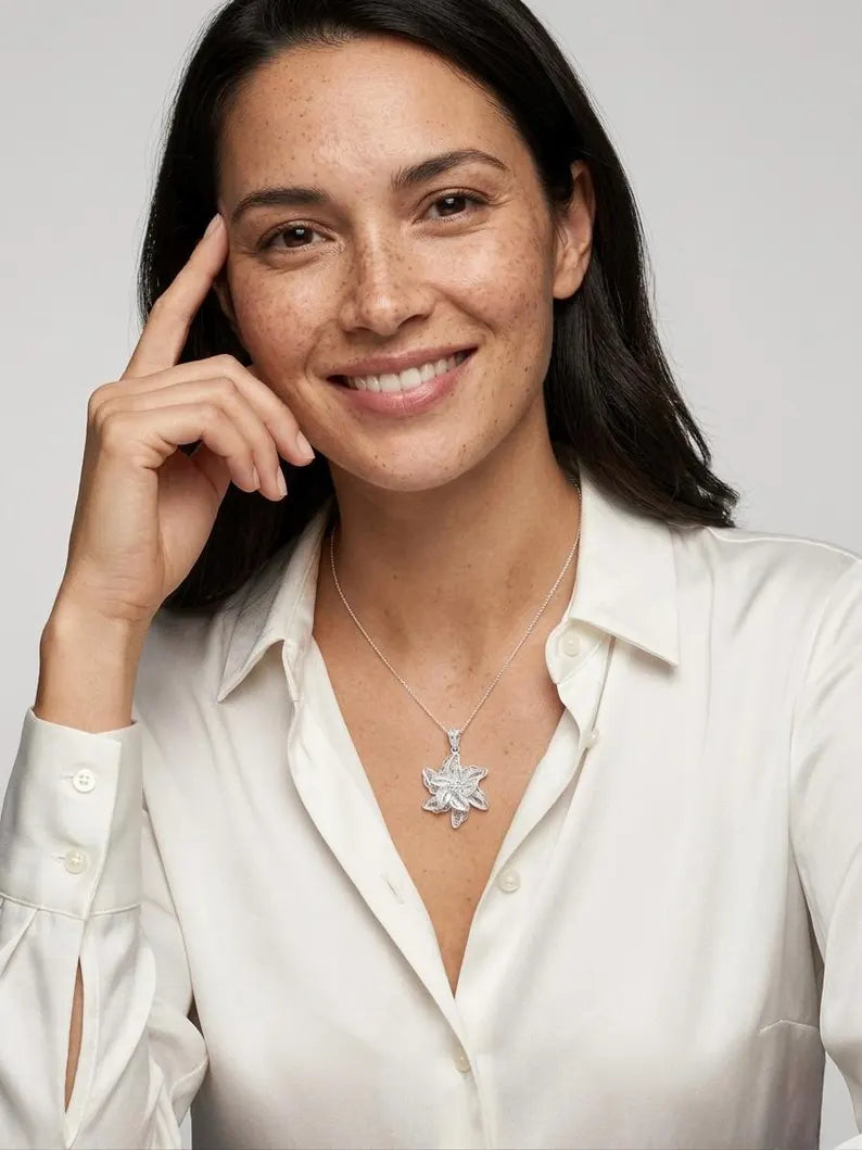 A smiling woman with dark hair wearing a white button-up shirt and a silver pendant necklace with a starburst or snowflake-like design, photographed against a light gray studio background.