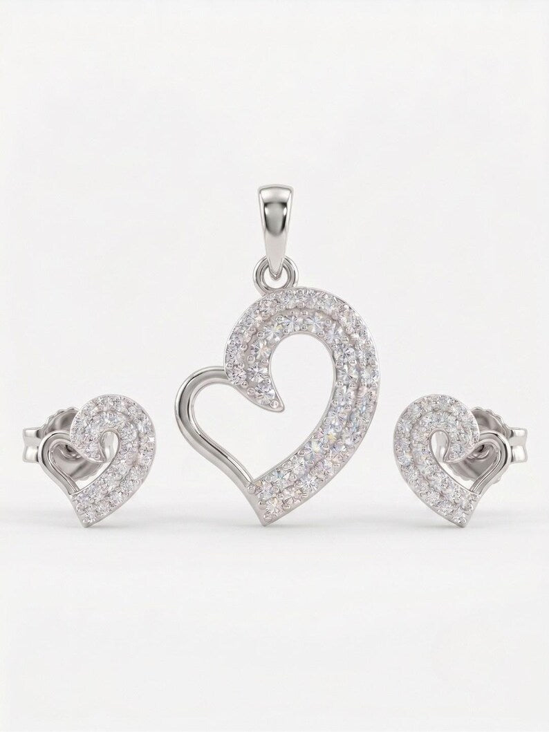 A silver heart-shaped jewelry set featuring a pendant, matching stud earrings, and a heart-shaped ring, all adorned with clear pavé gemstones on a white background.