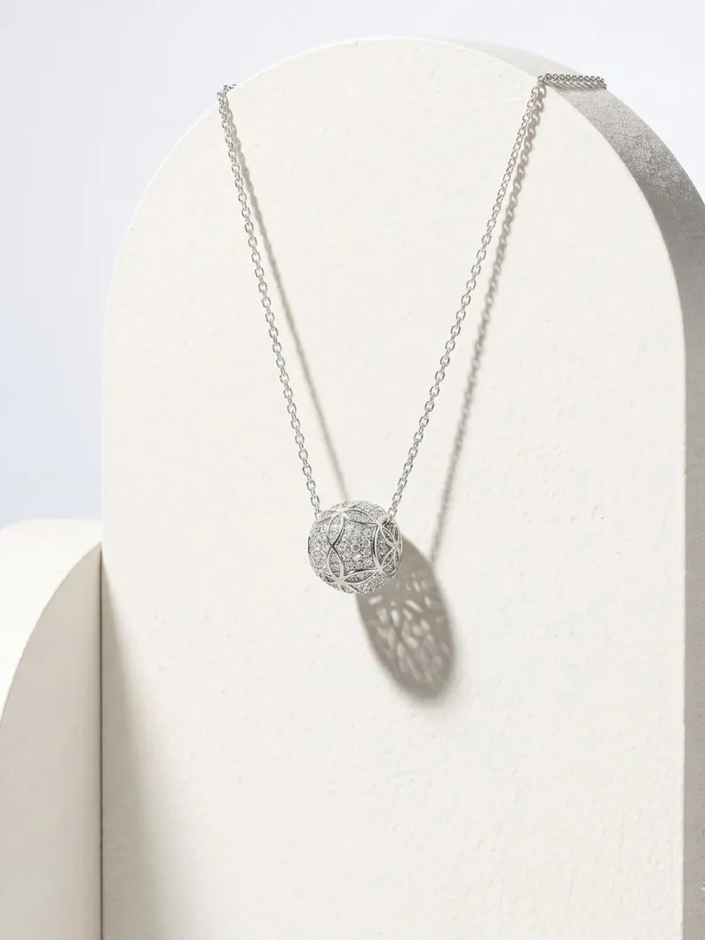 A silver filigree ball pendant necklace displayed on a textured white arch stand, casting a soft shadow on the right.