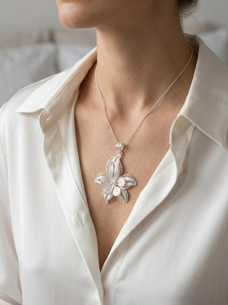 A close-up of a model wearing a silver floral pendant necklace on a delicate chain, styled with a white button-down shirt.