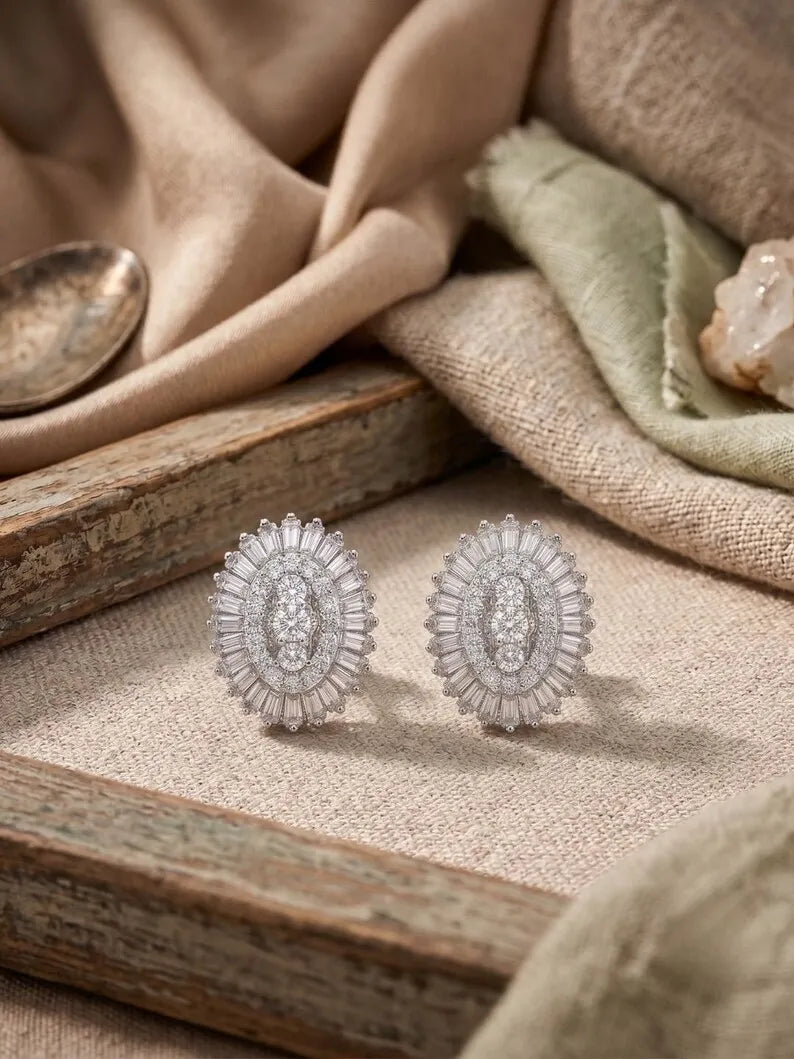 A pair of oval silver-toned earrings with radiating clear stones, displayed on a textured beige fabric surface with Oval halo stud earrings, rustic wood and soft draped textiles in the background.