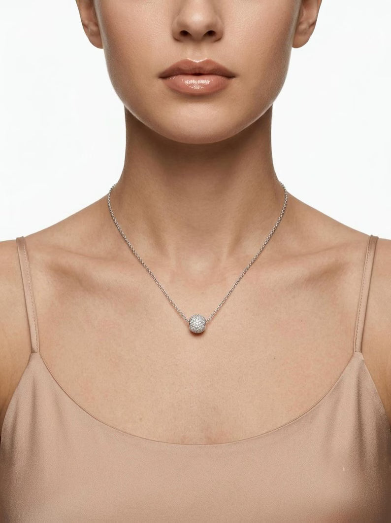 A close-up of a model wearing a delicate silver chain necklace with a small, round, diamond-like pendant, paired with a beige camisole top against a clean white background.