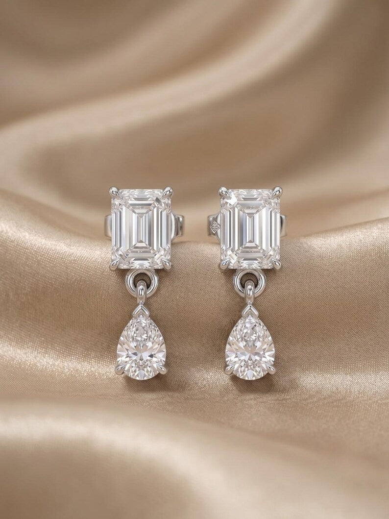 A pair of silver-toned drop earrings featuring a rectangular baguette-cut clear gemstone on the top and a pear-cut clear gemstone dangling below, displayed on a draped champagne satin fabric.