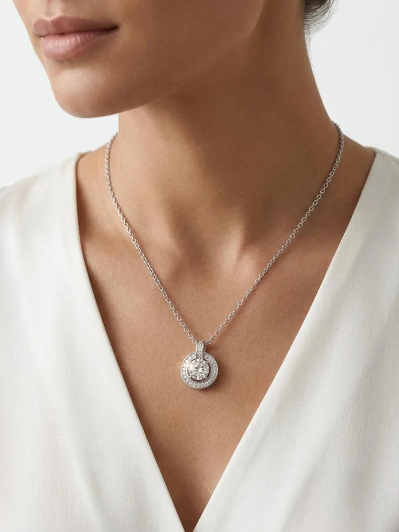 A close-up of a model wearing a delicate silver chain necklace with a round pendant set with clear gemstones, paired with a white V-neck top.