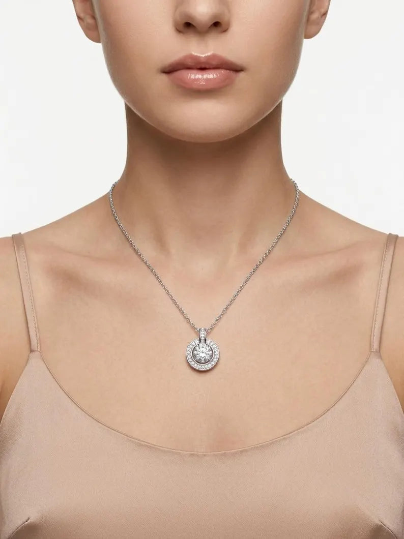 A close-up of a model wearing a silver pendant necklace with a round silver and clear-stone halo design, paired with a beige camisole top against a light gray background.