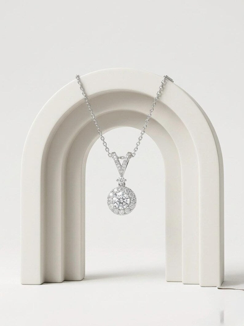 A silver-toned pendant necklace with a round pavé-style clear gemstone pendant, displayed on a white arched jewelry stand against a light gray studio background.