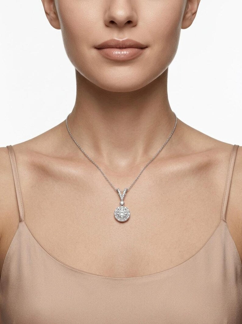 A close-up of a model wearing a delicate silver chain necklace with a round, clear-stone pendant on a light beige top against a white background.