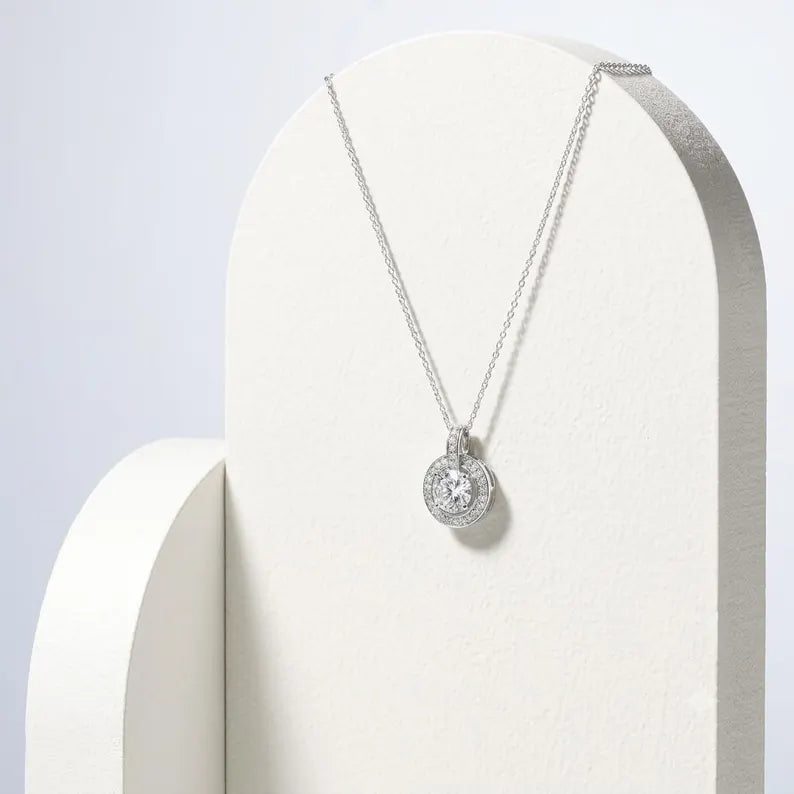 A silver-toned necklace with a round pendant displayed on a white arched jewelry stand against a light gray background.