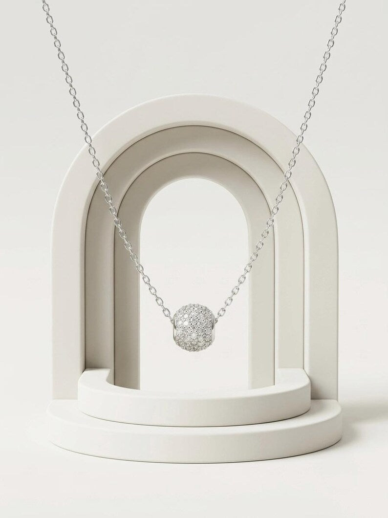 A silver chain necklace with a textured, diamond-like spherical pendant, displayed on a minimalist white geometric stand.