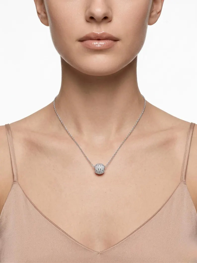 A close-up of a model wearing a delicate silver chain necklace with a small, round, clear gemstone pendant, paired with a beige camisole top against a white background.