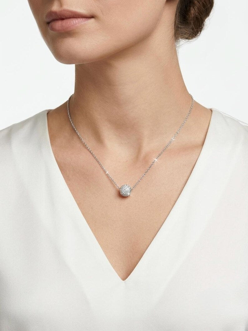 A close-up of a model wearing a delicate silver chain necklace with a round, clear pavé-set pendant, paired with a white V-neck top.