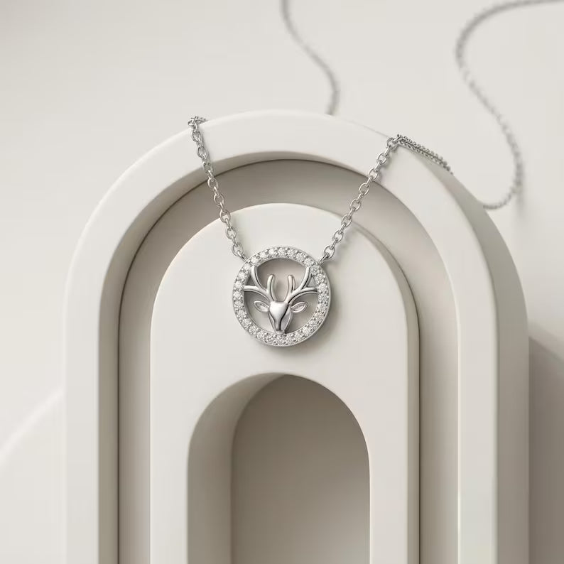 A silver-toned necklace with a circular pendant featuring a deer head motif, displayed on a sculptural white arch stand.