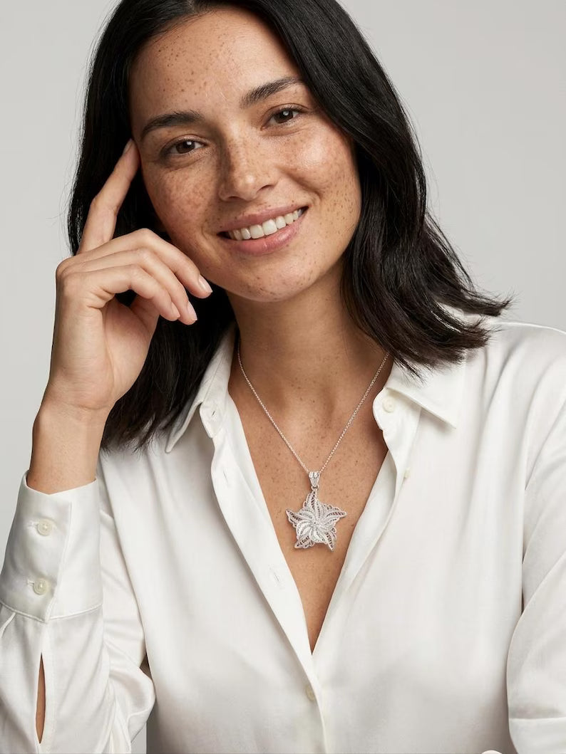 A smiling woman with dark hair wearing a white satin button-up shirt and a silver pendant necklace with a star-shaped floral design, set against a plain light gray background.