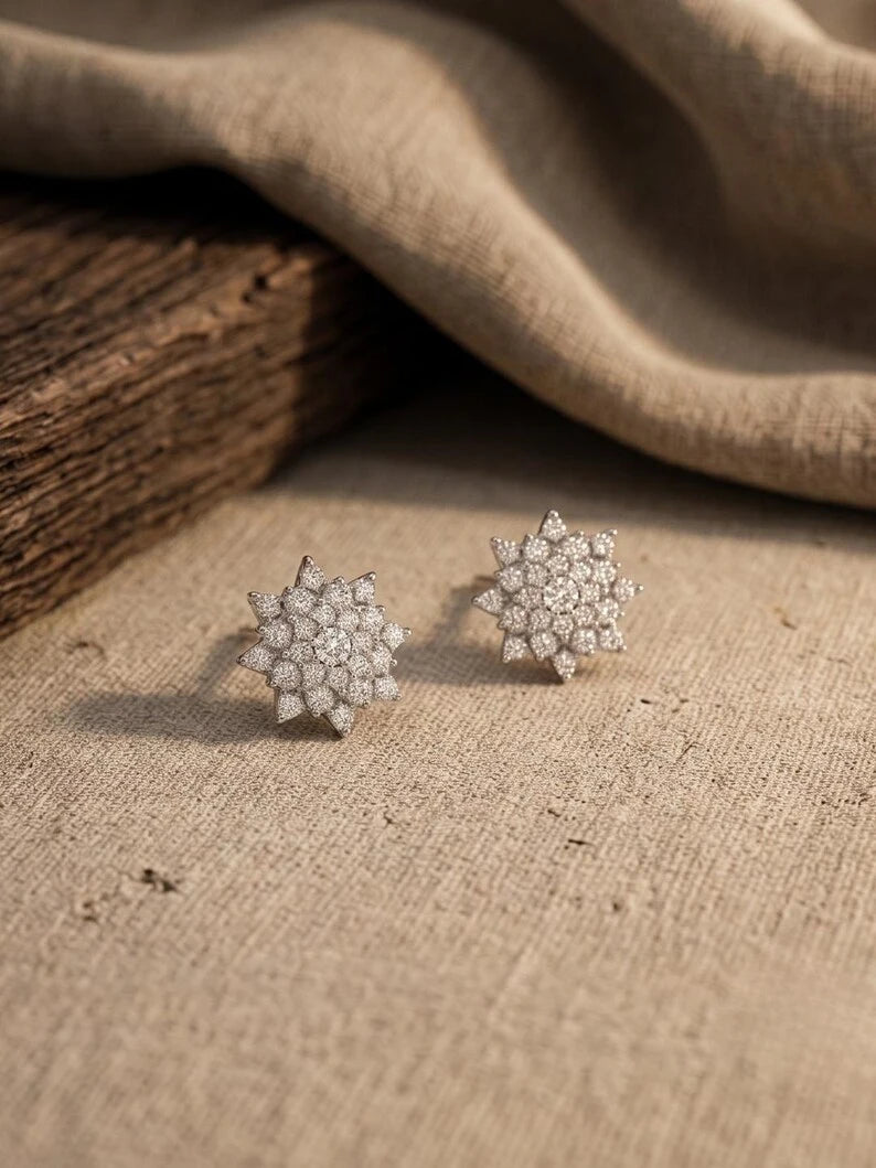 A pair of silver-toned snowflake stud earrings set with clear pavé gemstones, resting on a textured beige linen surface with a rustic wooden block and draped fabric in the background.