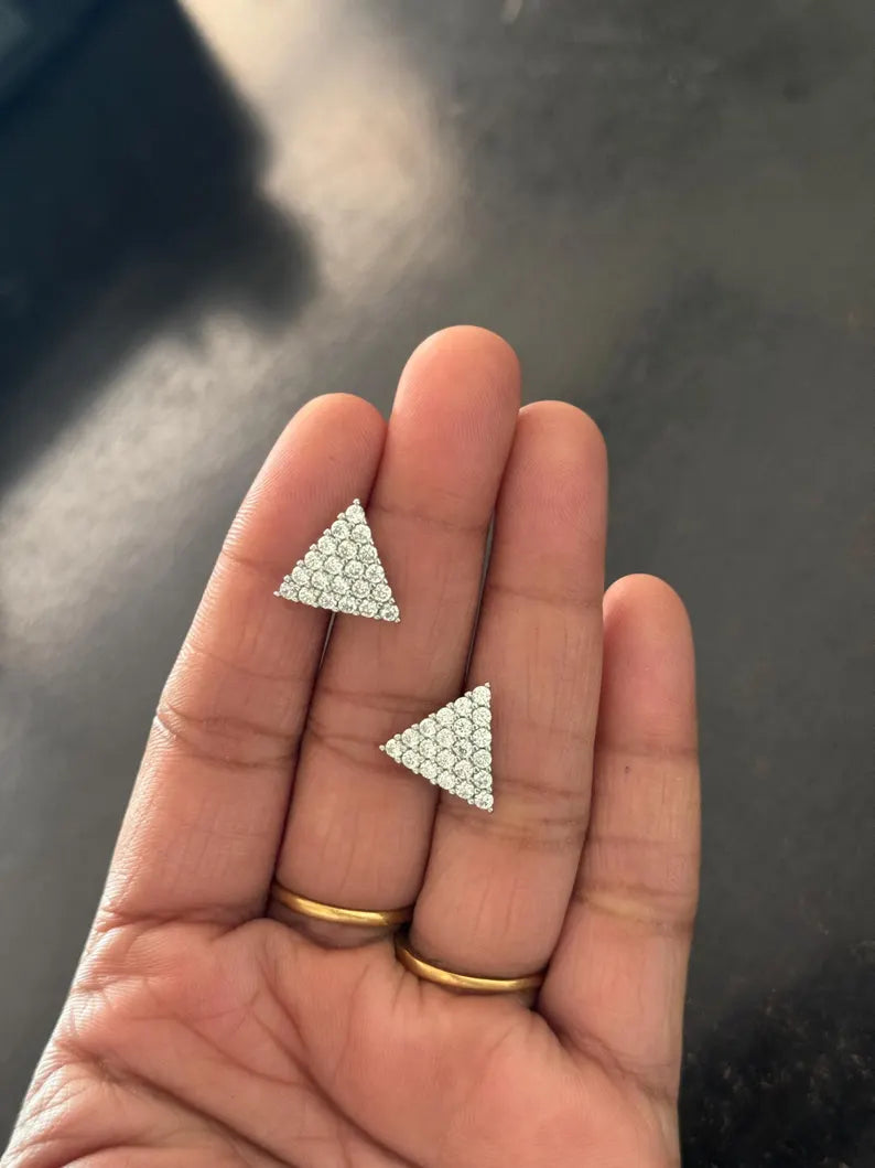 A close-up of a hand holding two small triangular stud earrings covered in clear rhinestones, with gold rings on the fingers against a dark gray background.