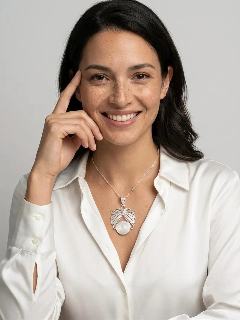 A smiling woman with dark hair wearing a white button-up shirt and a silver pendant necklace with a white pearl-like center, posed against a light gray studio background.