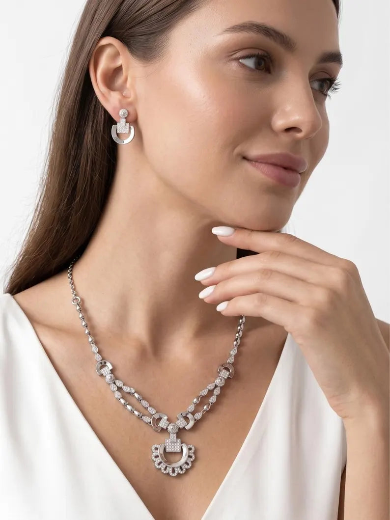 A close-up of a woman wearing a silver-toned double-strand necklace with a decorative crescent moon pendant and matching drop earrings, styled with a white top against a light gray background.