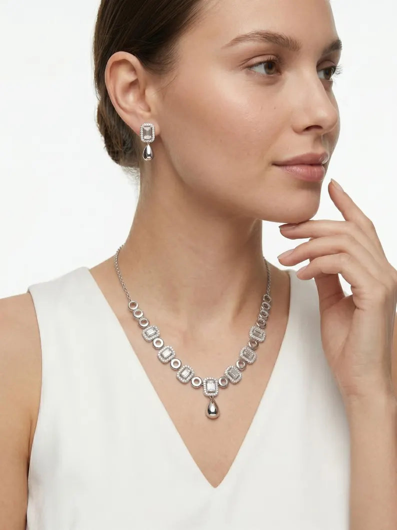 A woman wearing a silver-toned geometric diamond necklace and matching drop earrings, shown in a close-up portrait against a clean white background.
