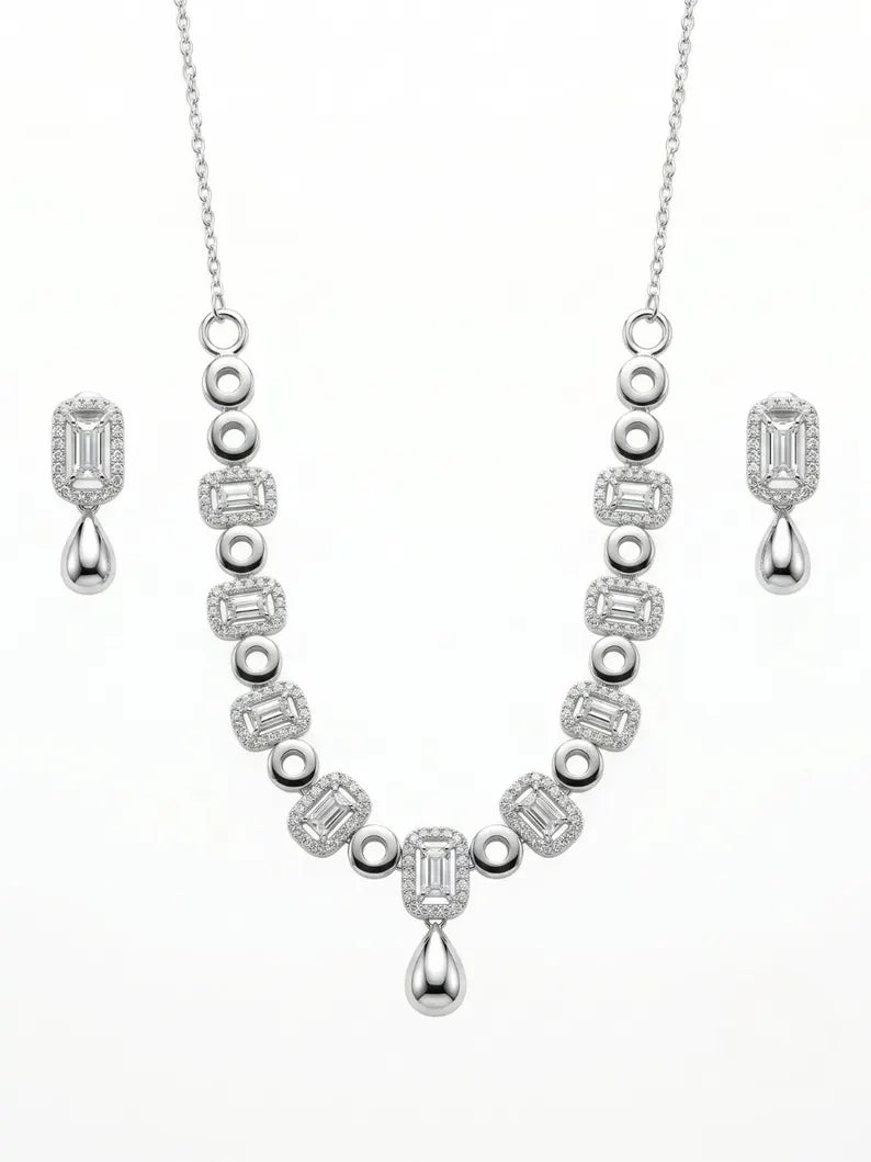 A silver-toned jewelry set featuring a necklace and matching drop earrings, both adorned with emerald-cut and round clear stones on a plain white background.