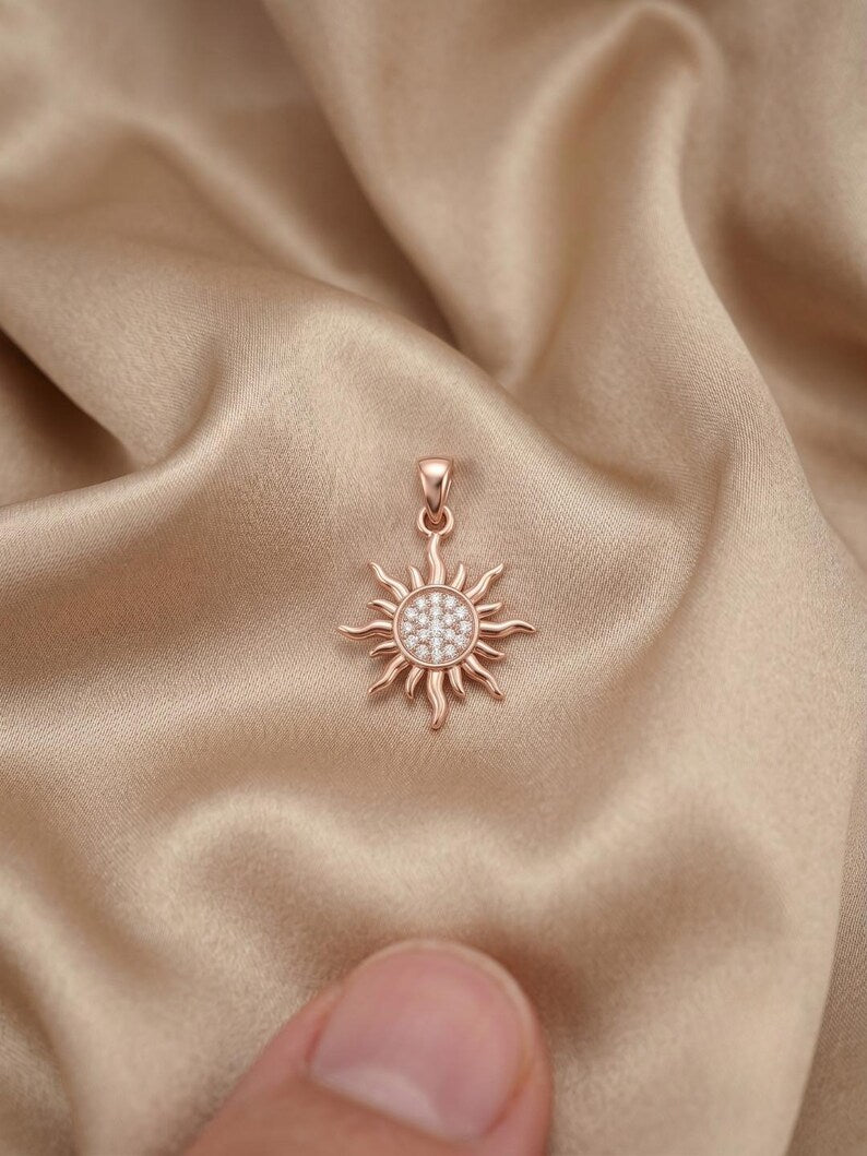 A rose gold sun pendant with a clear crystal center, resting on draped champagne-colored satin fabric with a finger visible at the bottom edge.