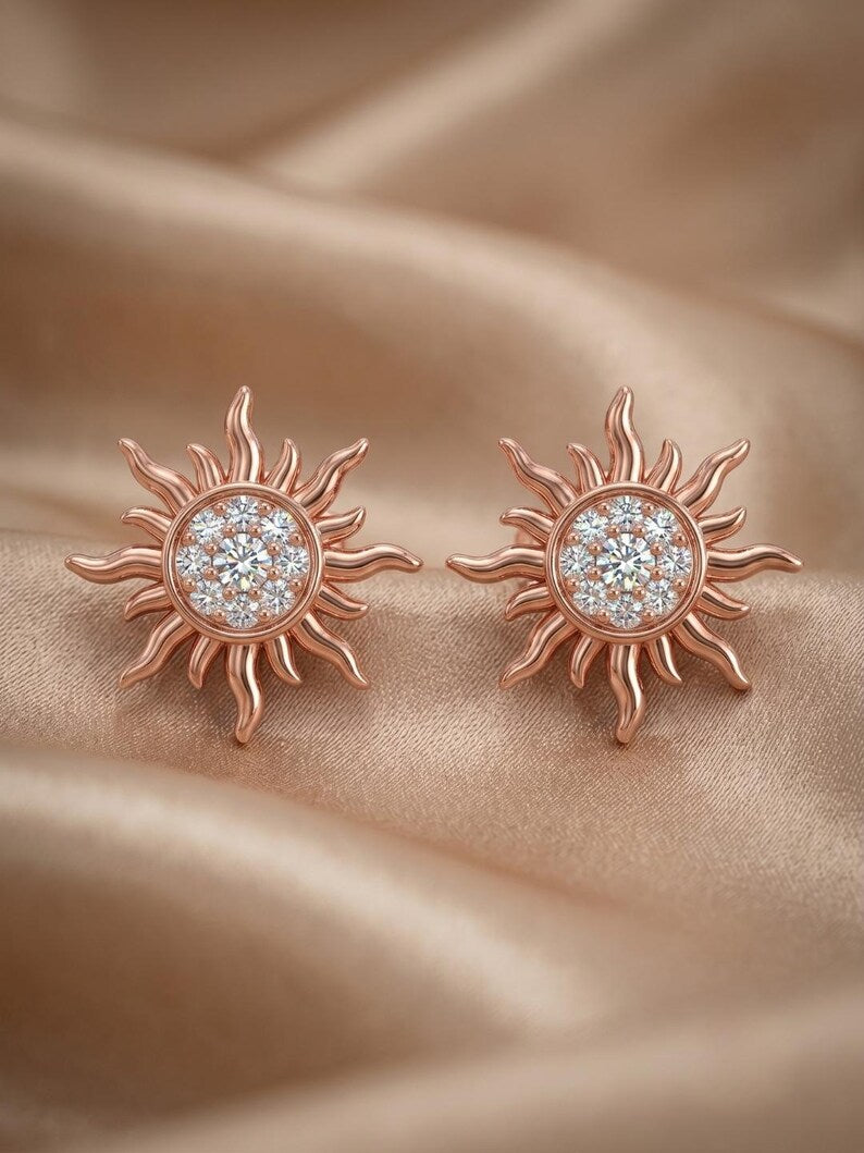 A pair of rose gold sun-shaped stud earrings with clear pavé gemstones in the center, displayed on a draped champagne-colored satin fabric.