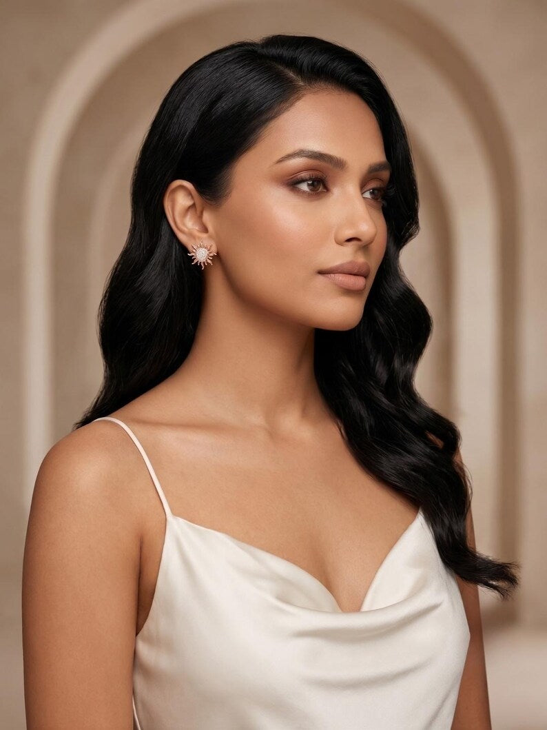 A woman with long, wavy black hair wearing a white spaghetti-strap top and a small, delicate starburst earring, posed against a warm beige background with an arched architectural detail.