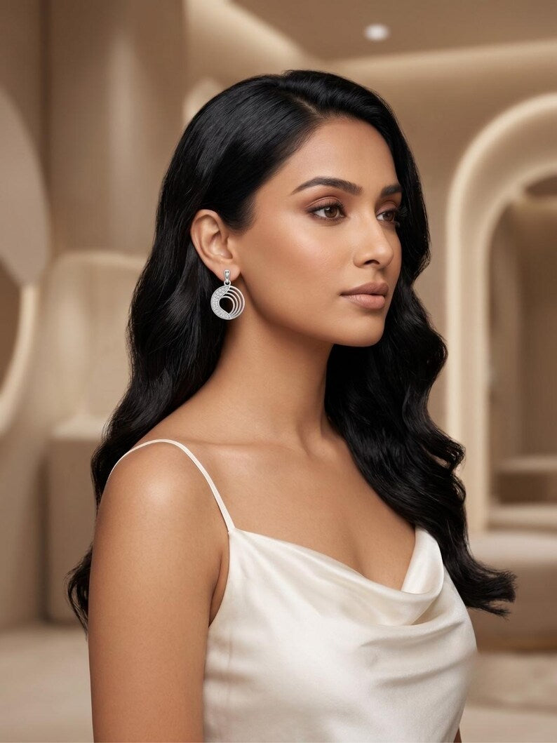 A woman with long, wavy black hair is shown in profile wearing a pair of silver-toned circular drop earrings with a small blue accent. She is wearing a white spaghetti-strap top against a softly lit, warm beige architectural background.