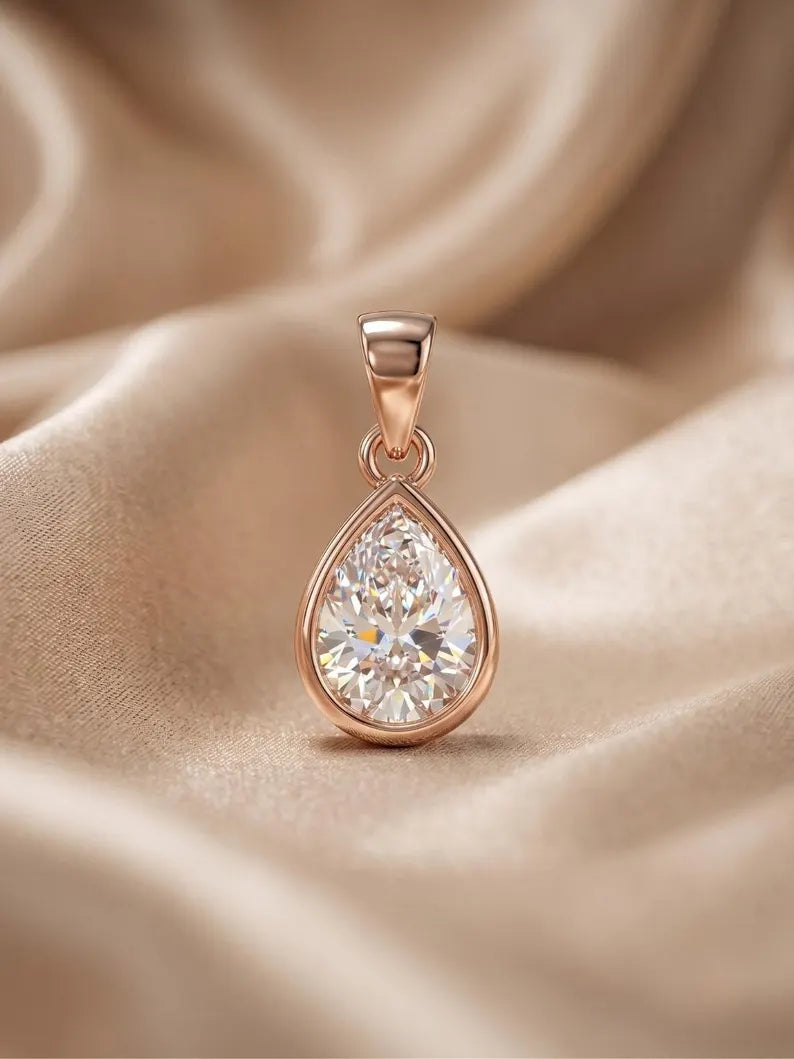 A teardrop-shaped pendant with a brilliant clear gemstone in a rose gold setting, resting on draped champagne-colored satin fabric.