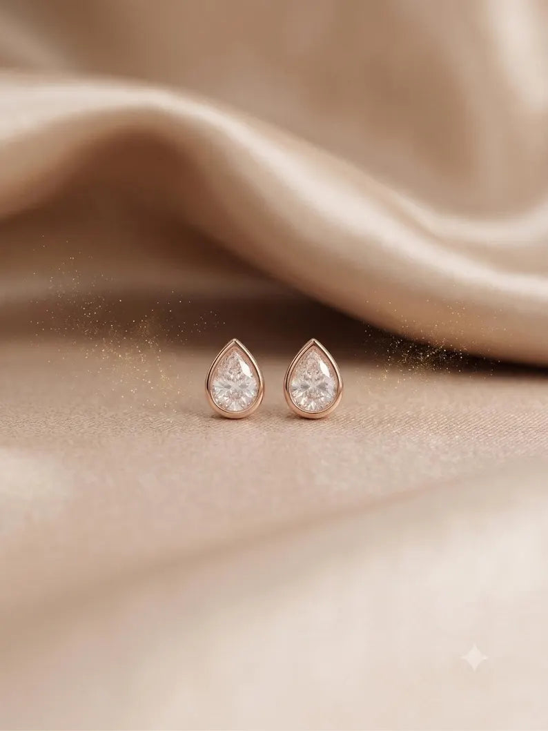 A pair of teardrop-shaped rose gold stud earrings with clear gemstones, displayed on a soft beige satin fabric background.