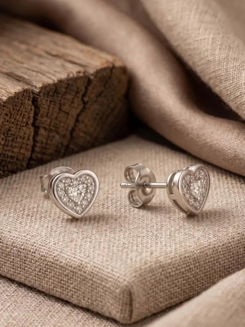 A pair of silver heart stud earrings with clear pavé stones, displayed on a textured beige fabric surface next to a rustic wooden block.