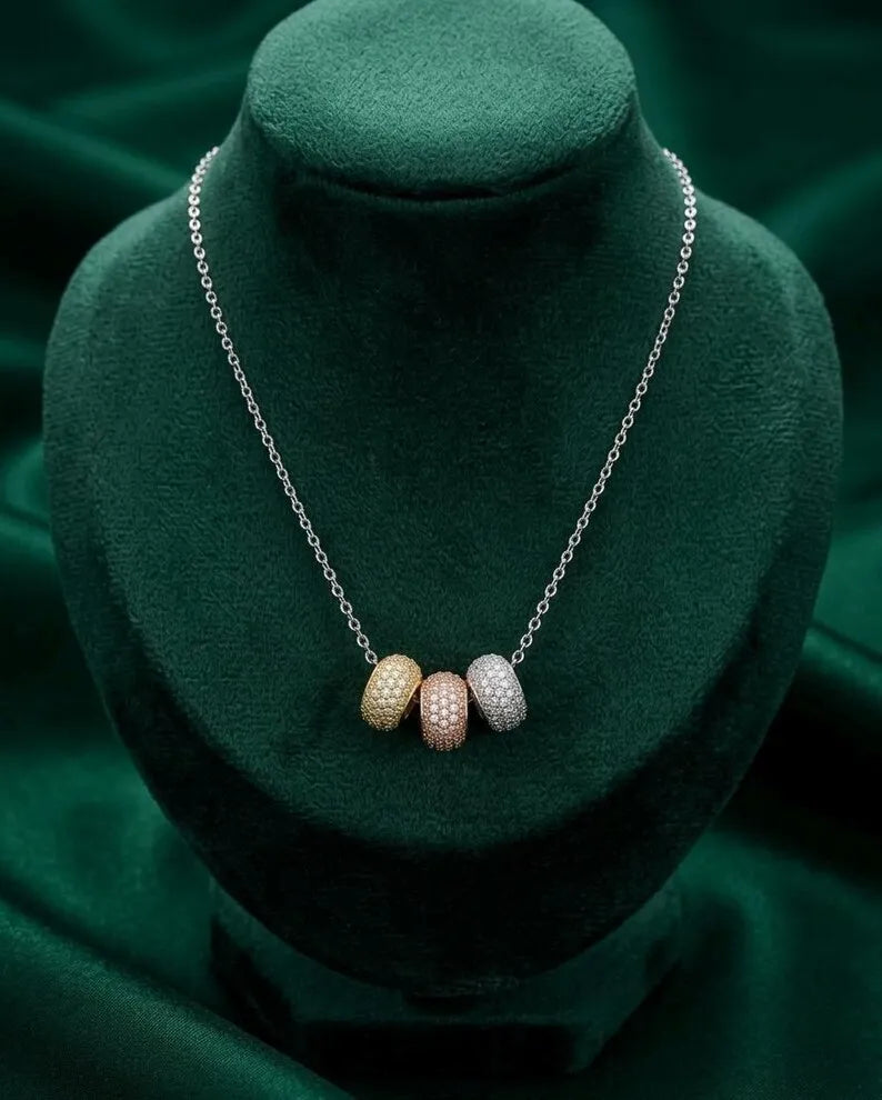 A silver chain necklace displayed on a dark green velvet jewelry bust, featuring three small oval pendant beads in gold, rose gold, and white textured settings.